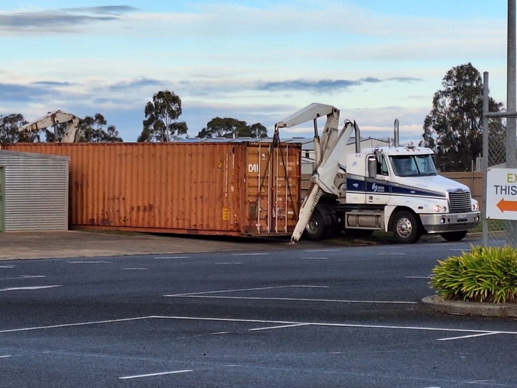 ID: Red shipping container being offloaded off a white truck in the church grounds.